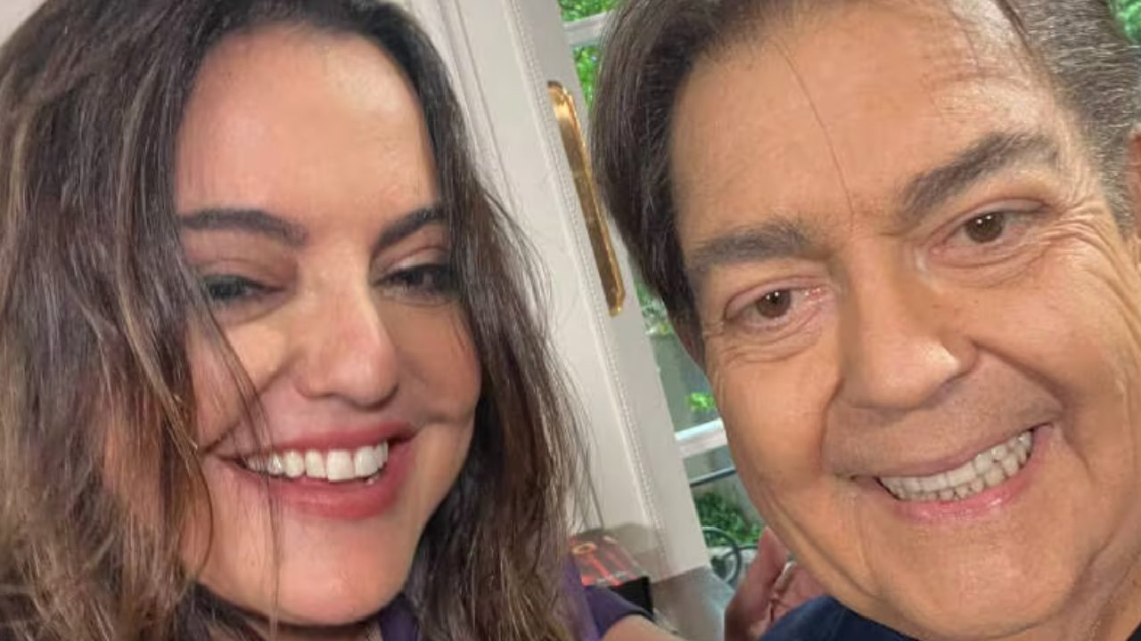 Selfie de um homem e uma mulher sorridentes em um ambiente interno bem iluminado, com uma janela e parte de uma maçaneta dourada visíveis ao fundo.