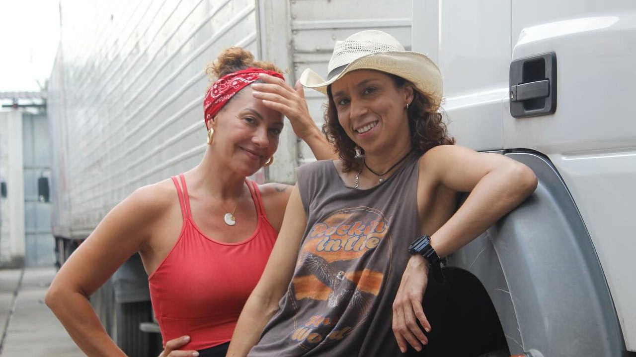 Fabiula Nascimento e Thalita Carauta posando sorridentes ao lado de um caminhão, com Fabiula usando blusa vermelha e lenço na cabeça e Thalita com chapéu de caubói e camiseta regata, em cena do piloto de Carga Pesada