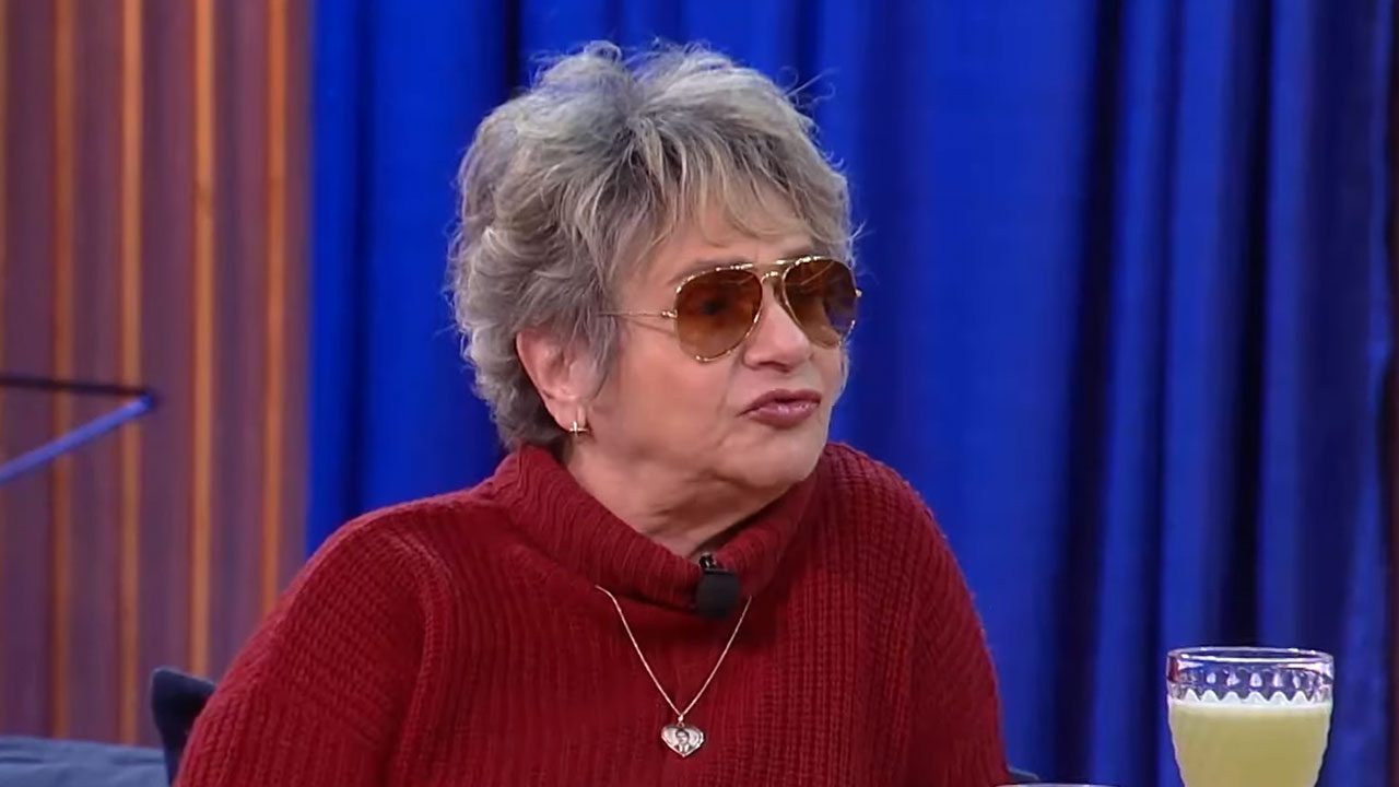 A atriz e comediante Fafy Siqueira com óculos de sol e suéter vermelho durante entrevista em programa com cenário de cortina azul