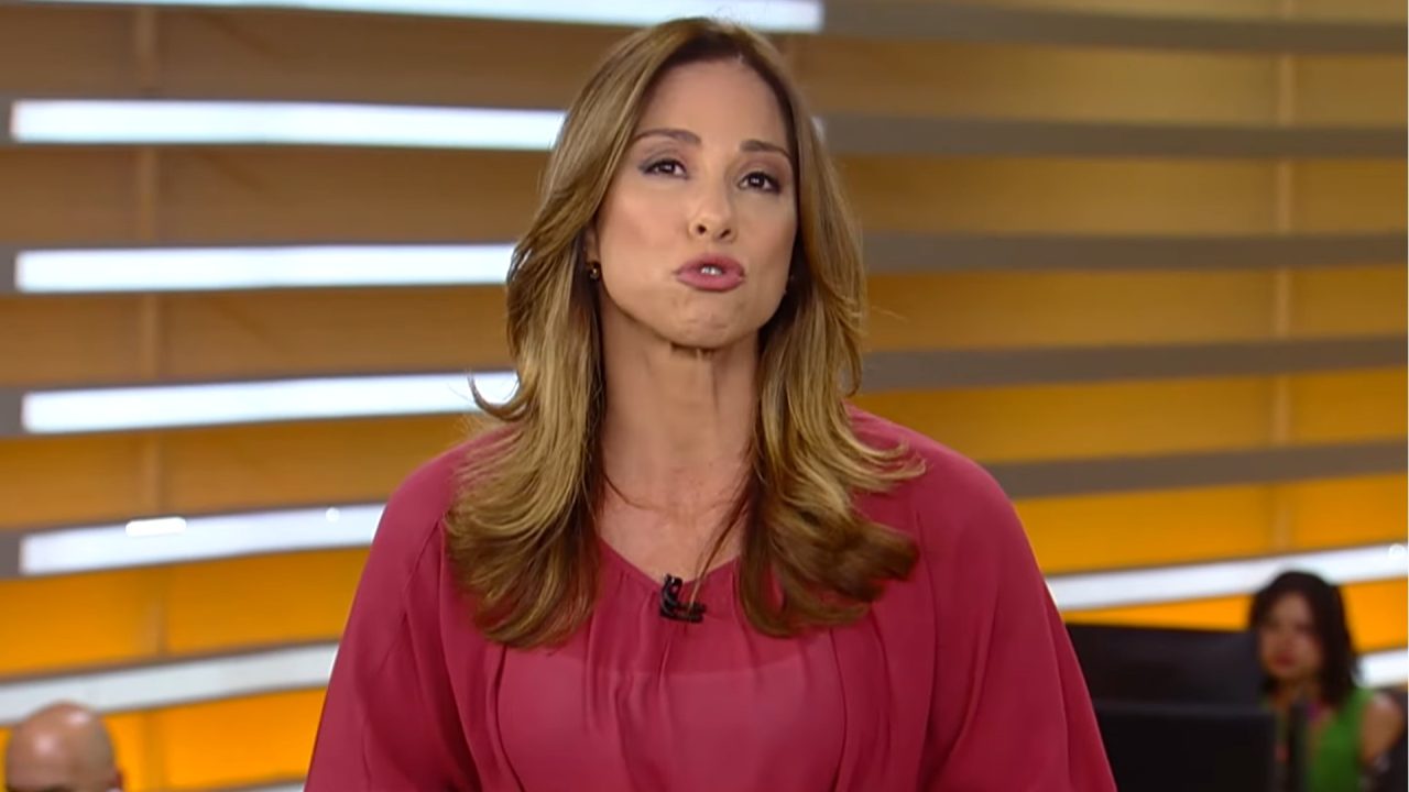 Neila Medeiros, uma mulher de cabelos castanhos e lisos apresenta o Fala Brasil em estúdio com fundo em tons de amarelo e laranja, vestindo blusa rosa e usando microfone preso à roupa