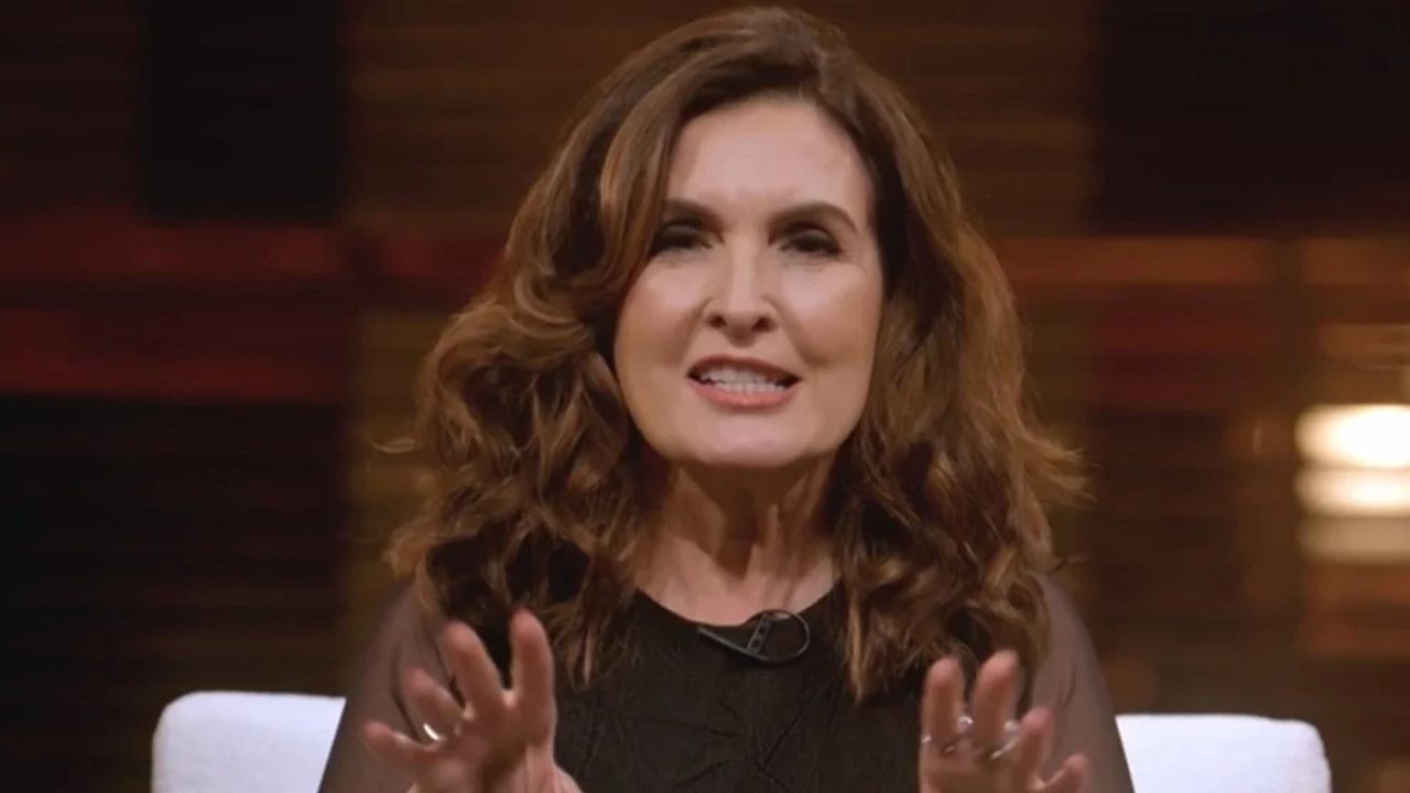 Mulher de cabelos castanhos ondulados, vestindo blusa preta, aparece sentada e falando com as mãos levemente erguidas; ao fundo, há iluminação quente e desfocada, criando um ambiente intimista de estúdio