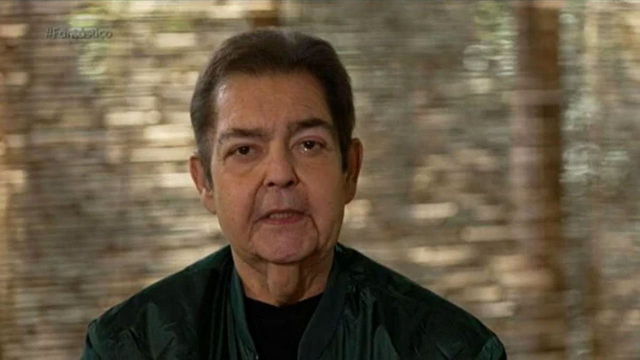Faustão fala diretamente para a câmera em gravação ao ar livre usando jaqueta verde e camiseta preta