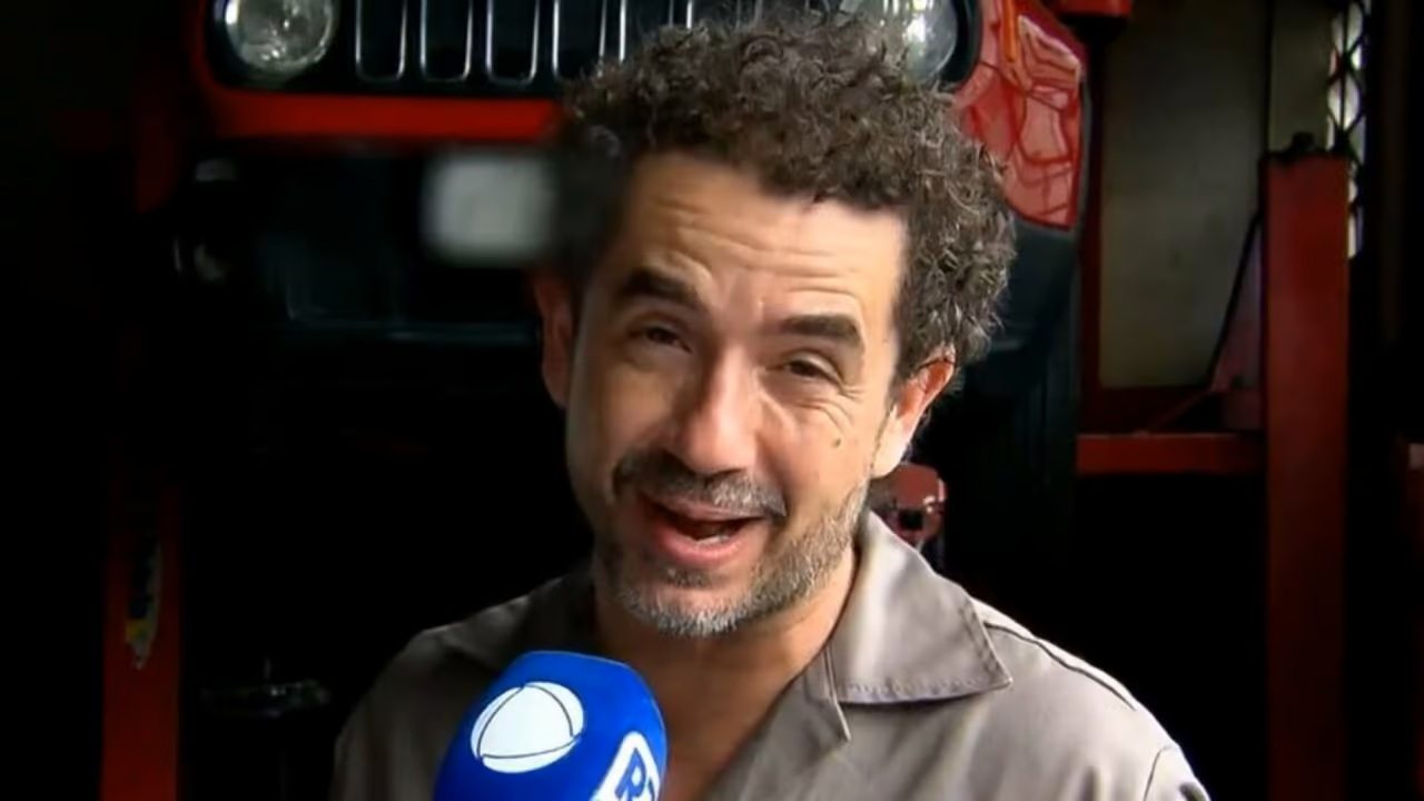 Homem com uniforme de trabalho sorri enquanto segura um microfone azul com o logotipo da Record TV, durante uma entrevista em uma oficina mecânica com um carro vermelho elevado ao fundo