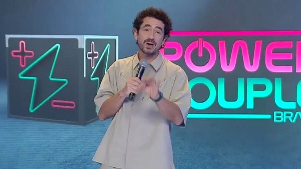 Felipe Andreoli em um estúdio, segurando um microfone e falando diretamente para a câmera. Ele usa roupas claras e tem cabelo curto e levemente cacheado, com barba discreta. Ao fundo, há um painel com elementos gráficos em neon, incluindo um símbolo de energia e a palavra “Power Couple Brasil”, indicando um programa de televisão. O ambiente é iluminado e moderno, típico de apresentação ou entretenimento televisivo.