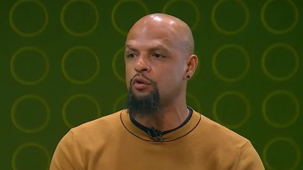 A imagem mostra um homem careca com barba e cavanhaque, usando brinco e uma blusa marrom clara. Ele fala enquanto está em um estúdio de televisão, com fundo verde estampado com círculos em tom mais escuro, sugerindo um programa esportivo ou de entrevistas