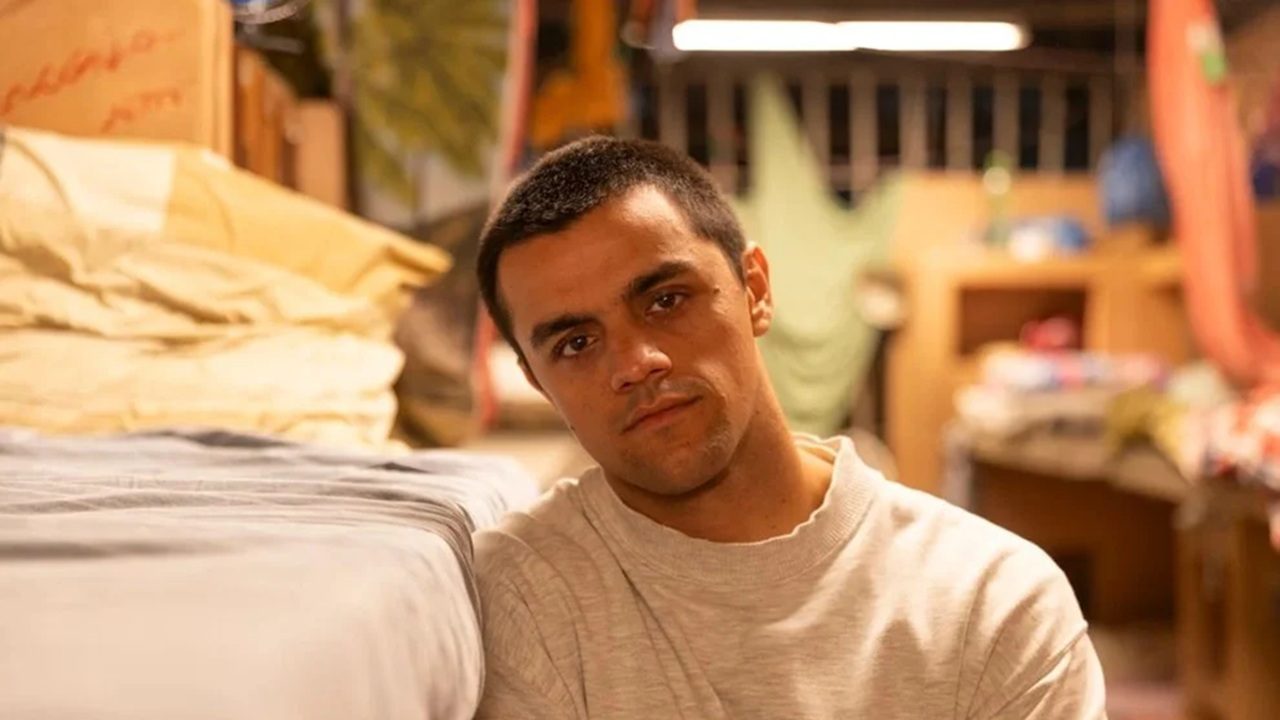 Homem sentado ao lado de beliche olha sério em quarto simples iluminado por luz quente