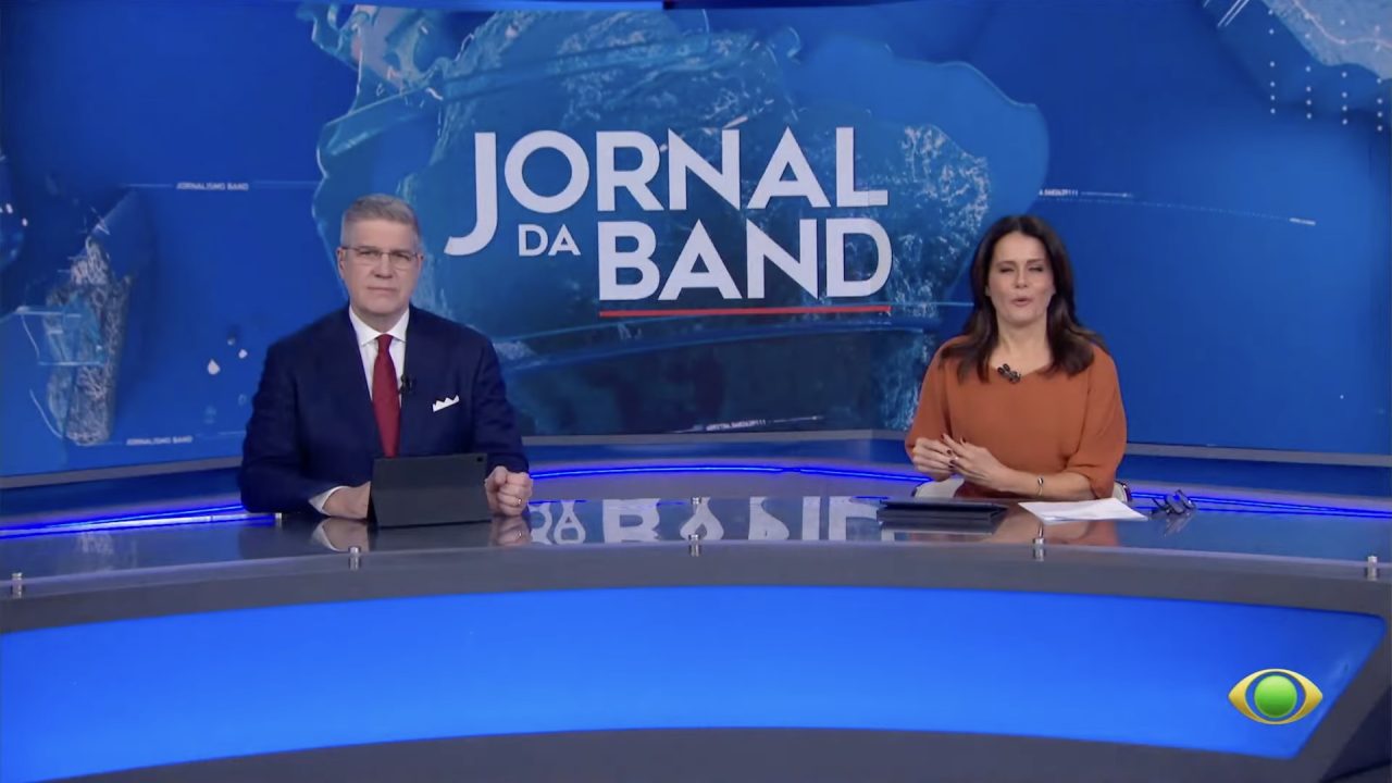 Felipe Vieira, de terno e gravata, e Adriana Araújo, de blusa laranja, na bancada do Jornal da Band, com telão do programa ao fundo