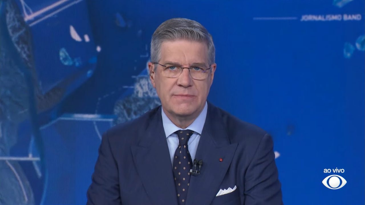 Felipe Vieira usa terno escuro e gravata com pontos enquanto apresenta o Jornal da Band em estúdio com fundo azul e gráficos digitais; no canto da tela aparece a indicação ao vivo e o logotipo da Band.