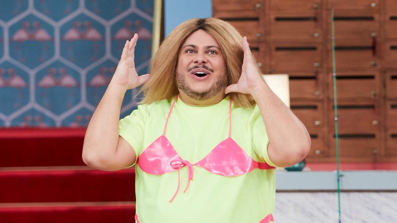 Ferdinando de Marcus Majella levanta as mãos e sorri com peruca loira e top rosa em cena de Vai Que Cola