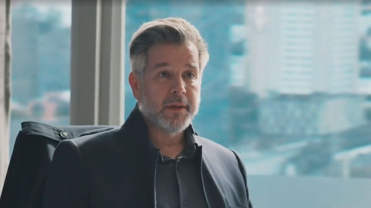 Homem de meia-idade com barba grisalha e cabelos penteados para o lado aparece em um escritório iluminado, usando casaco escuro sobre camisa cinza. Ao fundo, vê-se uma grande janela com a paisagem urbana desfocada, composta por prédios e ruas