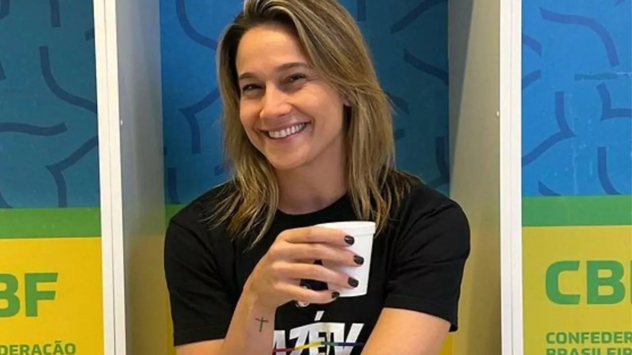 Mulher com camiseta preta sorri segurando copo em frente a painéis da CBF com fundo azul e amarelo