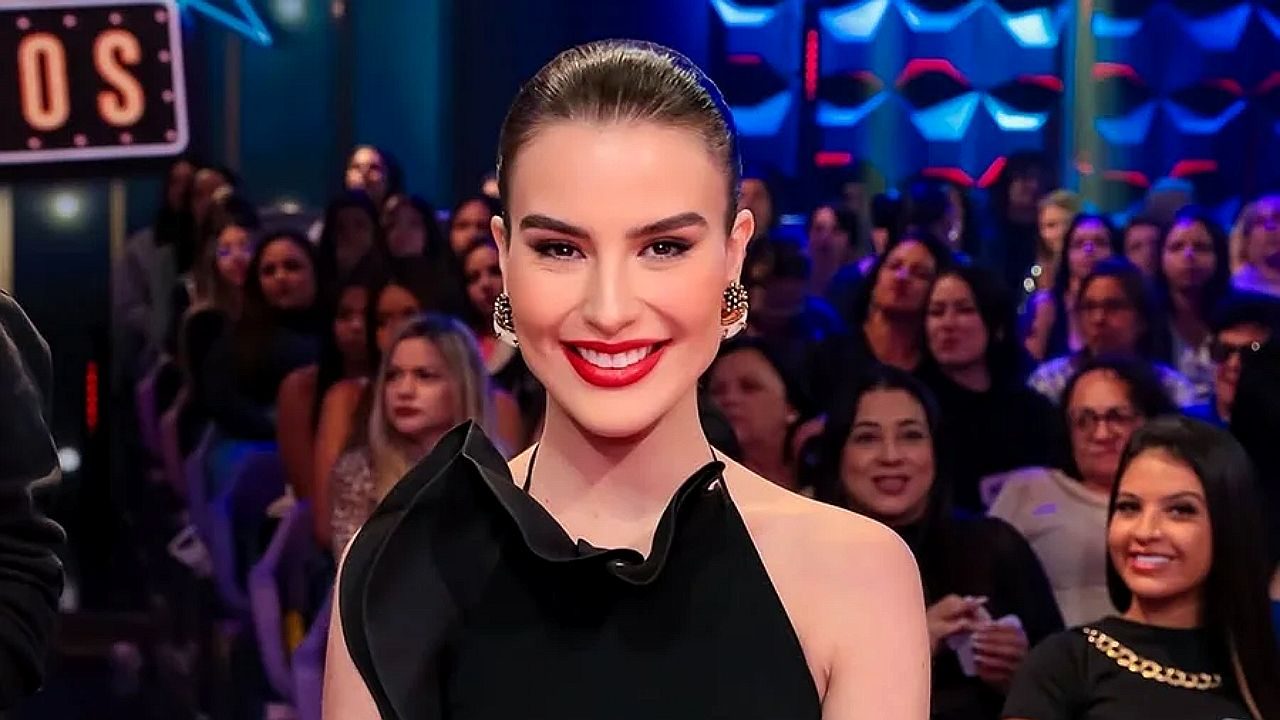 Apresentadora de vestido preto sorri em programa de auditório com plateia ao fundo