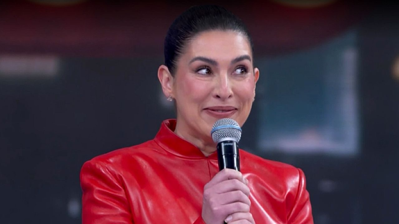 Apresentadora com roupa vermelha segura microfone e sorri durante programa de TV em estúdio