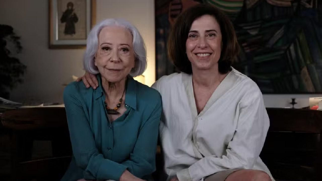 Fernanda Montenegro e Fernanda Torres sentadas lado a lado, sorrindo para a câmera em um ambiente interno. A mulher à esquerda tem cabelos grisalhos e usa uma blusa azul; a da direita tem cabelos castanhos curtos e veste uma camisa clara