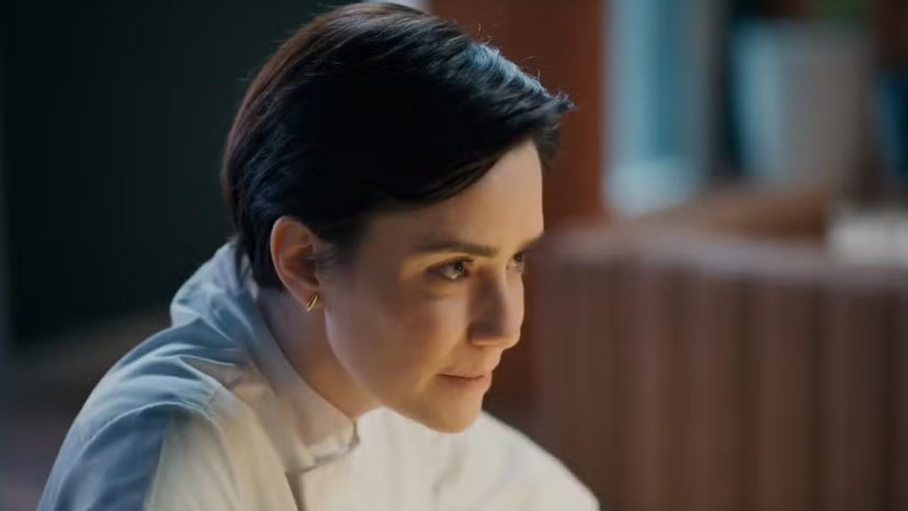 Fernanda Vasconcellos de cabelo curto e escuro veste camisa branca e observa algo com expressão concentrada em ambiente interno iluminado por luz natural