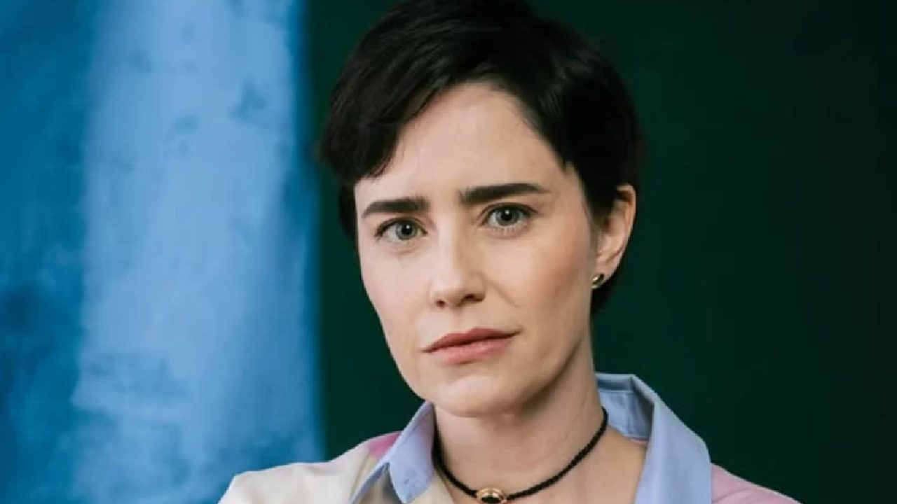 Fernanda Vasconcellos em retrato posado, com cabelo curto e expressão séria, usando camisa em tons claros e colar preto com pingente dourado, diante de fundo azul e verde