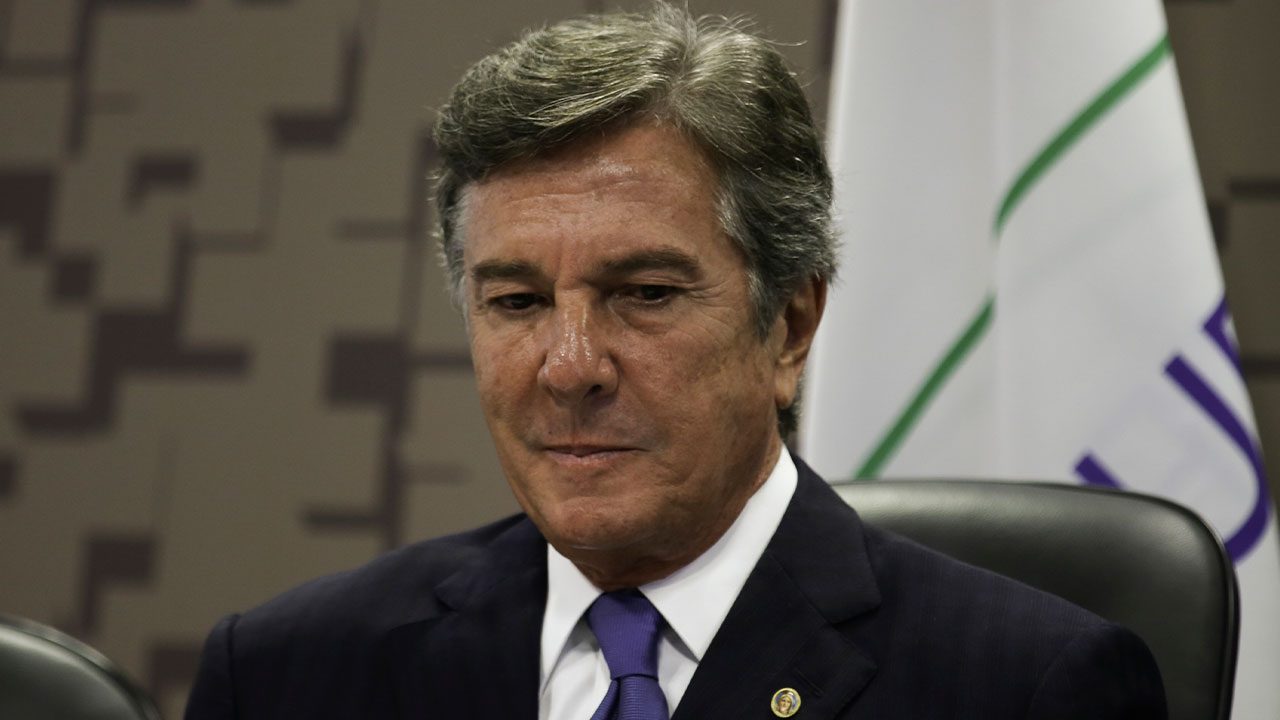 Fernando Collor de terno azul-marinho e gravata roxa em close-up com expressão séria em ambiente formal