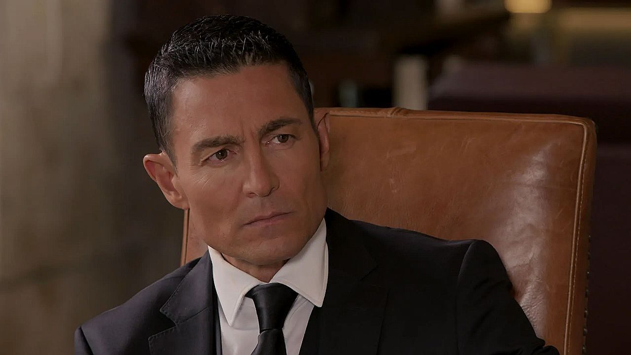 Homem de terno preto e gravata está sentado em poltrona de couro marrom, com expressão séria e olhar desconfiado voltado para alguém fora de cena. O ambiente interno elegante e iluminação suave sugerem cena dramática em contexto corporativo ou familiar.