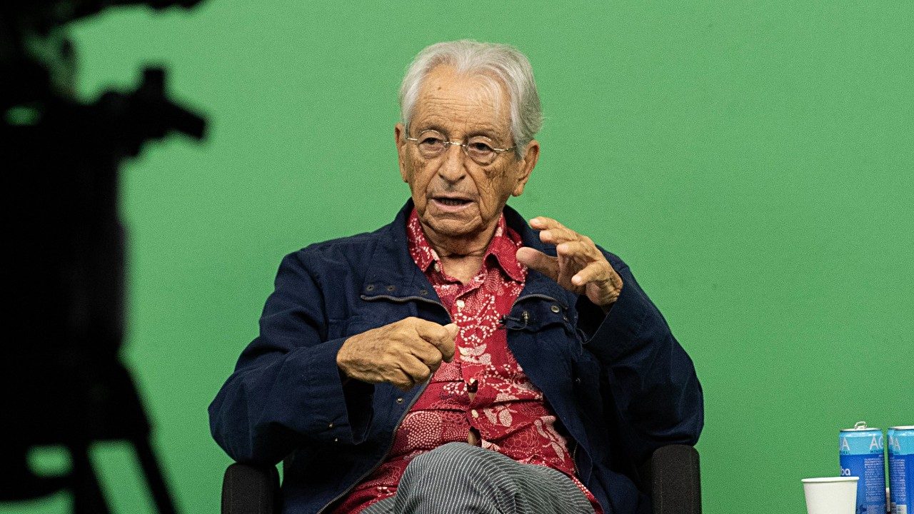 Fernando Gabeira fala gesticulando com as mãos sentado em cadeira durante evento com fundo verde e garrafa de água ao lado