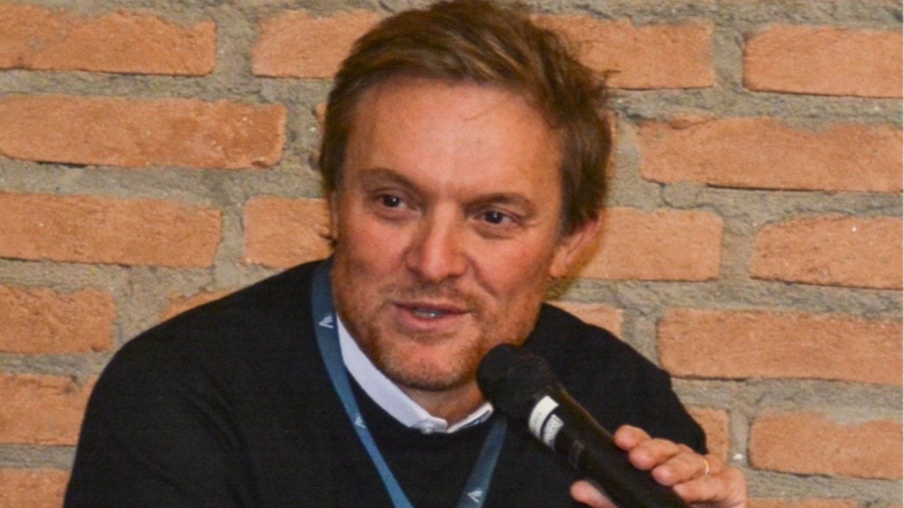 Fernando Stucchi falando ao microfone durante evento diante de parede de tijolos aparentes
