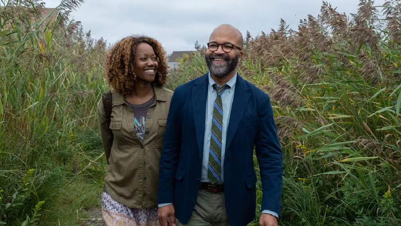 Casal caminha sorridente por uma trilha cercada por vegetação alta; mulher usa jaqueta verde e saia estampada, enquanto homem veste paletó azul, gravata listrada e óculos, em clima leve e descontraído