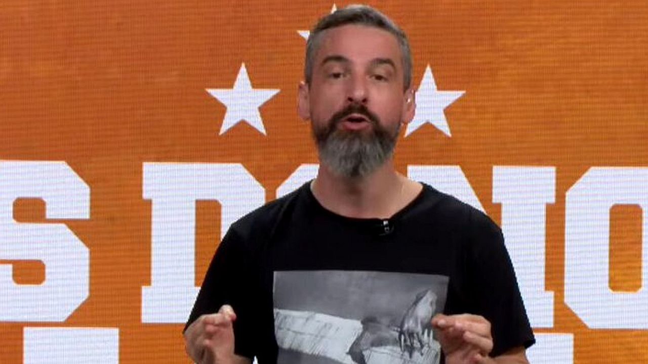 Apresentador barbudo fala em frente a telão laranja com estrelas durante programa de TV