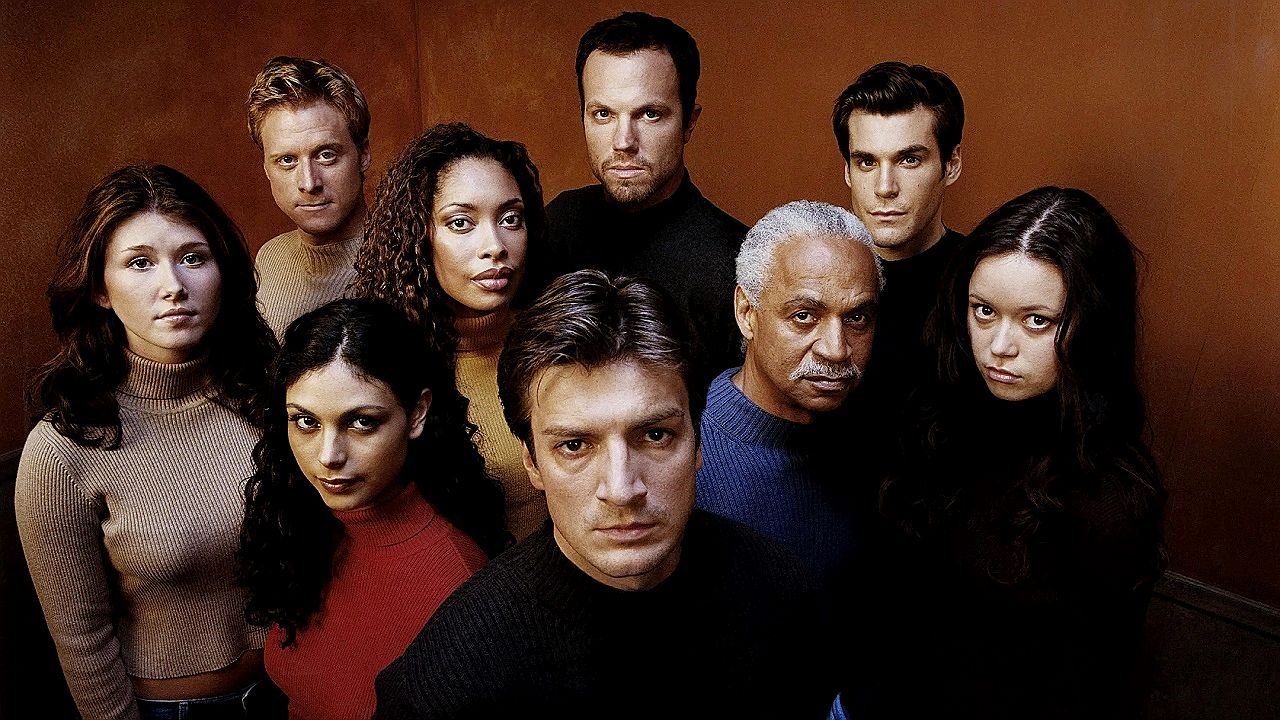 Um grupo de nove pessoas está posando junto diante de um fundo marrom simples. Todos olham diretamente para a câmera com expressões sérias ou intensas. Eles estão posicionados em diferentes alturas, formando duas fileiras, com um homem ao centro em primeiro plano. As roupas são em tons neutros, como preto, marrom e bege, incluindo suéteres e blusas de gola alta. A composição da imagem sugere um retrato promocional de elenco, com iluminação uniforme destacando os rostos de cada personagem.