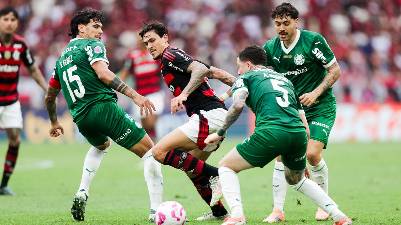 Quatro jogadores em campo, um do Flamengo com camisa preta e vermelha, e três do Palmeiras com camisa verde, disputando a bola