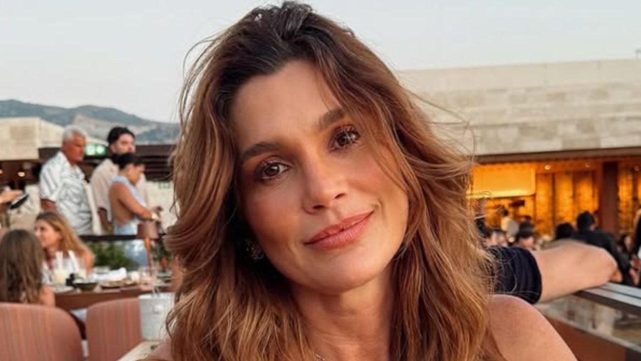 Mulher de cabelos castanhos soltos sorri levemente em ambiente externo com pessoas ao fundo