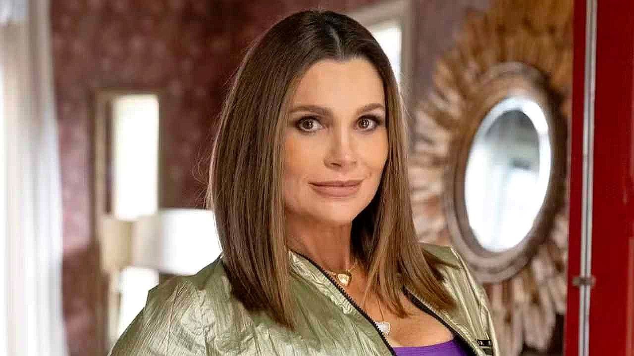 Atriz com jaqueta verde posa em ambiente interno com espelho ao fundo em cena de novela