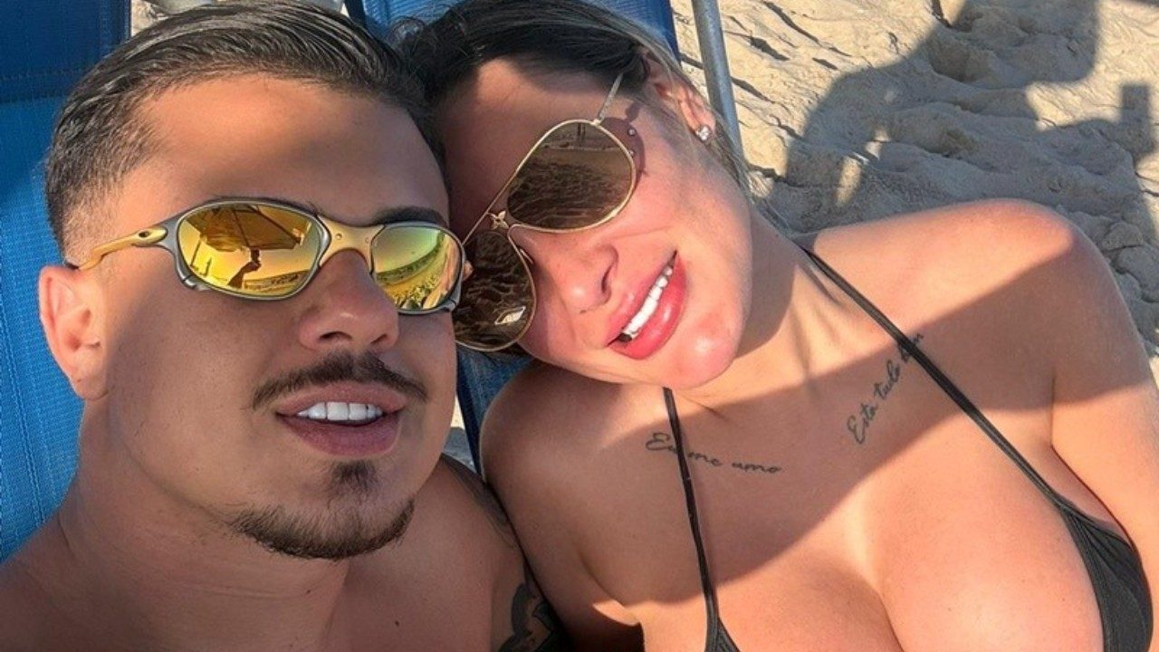Flávio Giglioli e Andressa Urach deitados em cadeiras de praia usando óculos escuros enquanto posam juntos na areia com mar ao fundo