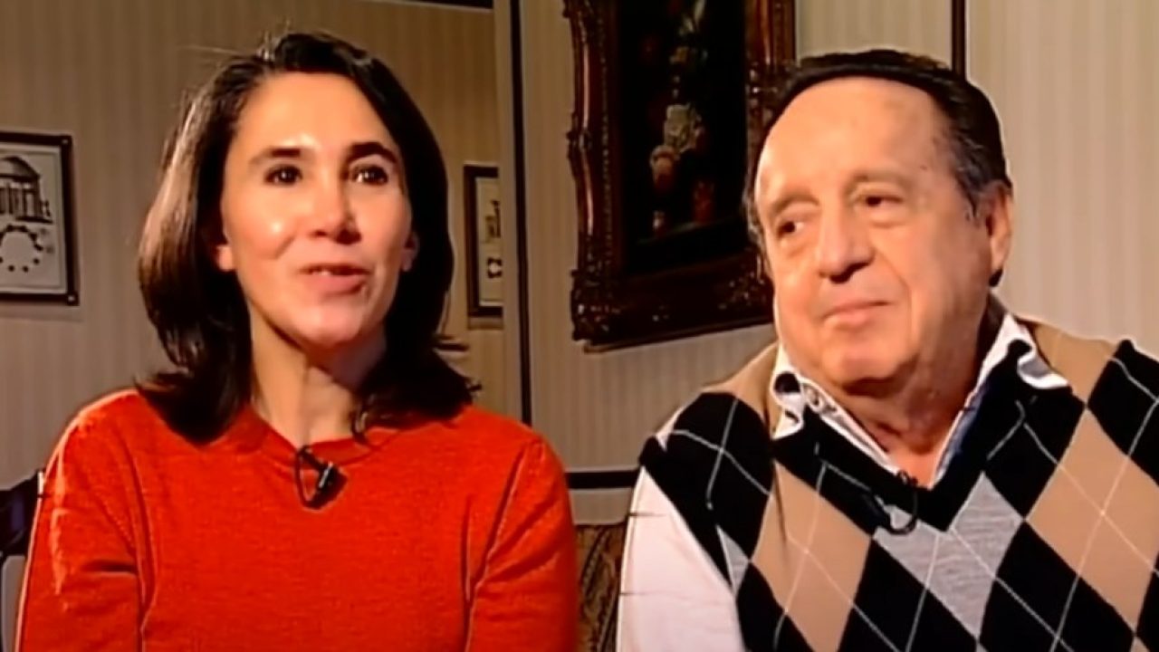 Duas pessoas sentadas lado a lado em um ambiente interno, durante uma entrevista. À esquerda, uma mulher de meia-idade veste blusa vermelha e olha levemente para a frente, com expressão atenta e serena. À direita, um homem mais velho usa camisa clara com colete de losangos em tons neutros e observa a mulher com um leve sorriso. Ao fundo, há quadros na parede e iluminação quente, sugerindo uma sala de estar ou ambiente doméstico acolhedor.