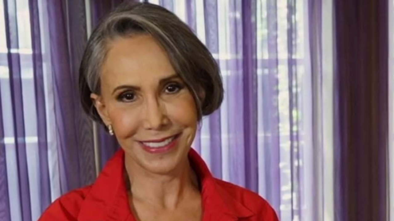 Mulher madura sorri em ambiente interno iluminado por luz natural. Ela tem cabelos curtos grisalhos penteados de lado e veste camisa vermelha. Ao fundo, cortinas lilás semi-transparentes deixam entrar a claridade