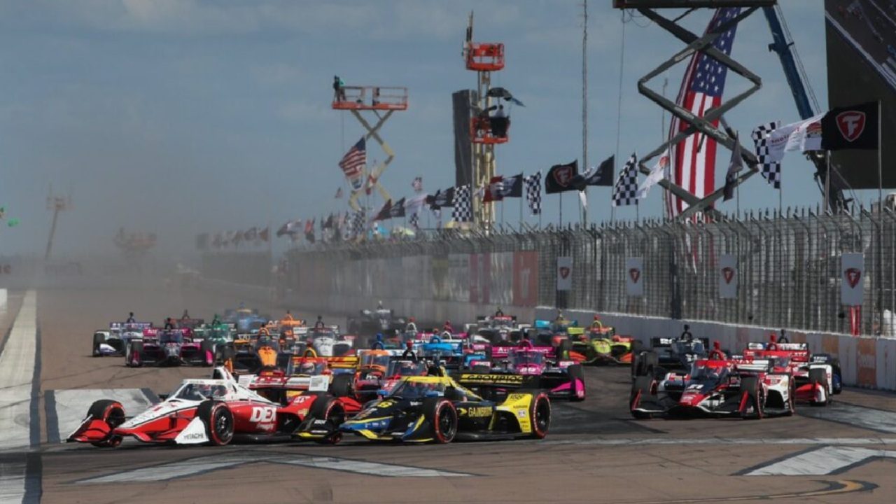 Carros alinham-se na largada de corrida da IndyCar com bandeiras quadriculadas ao fundo