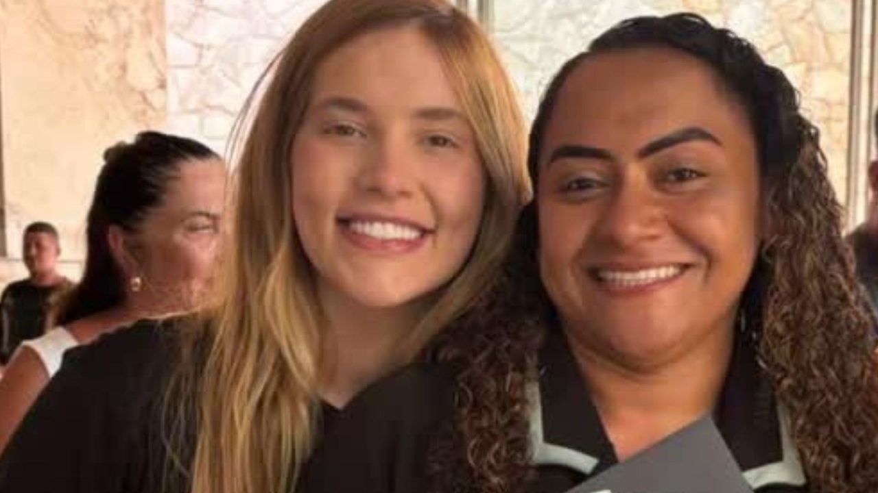 Duas mulheres sorridentes posam lado a lado em ambiente iluminado com outras pessoas ao fundo