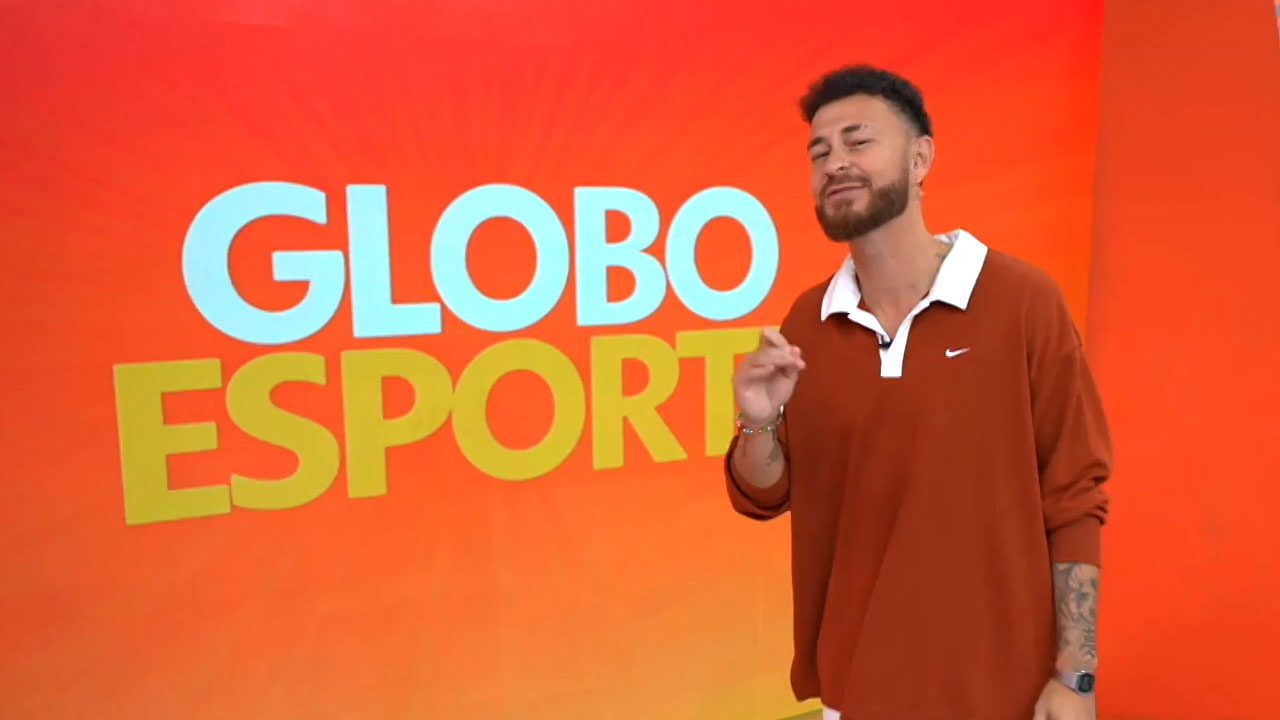 Fred Bruno em frente ao telão do Globo Esporte usando camisa polo larga marrom com detalhes brancos