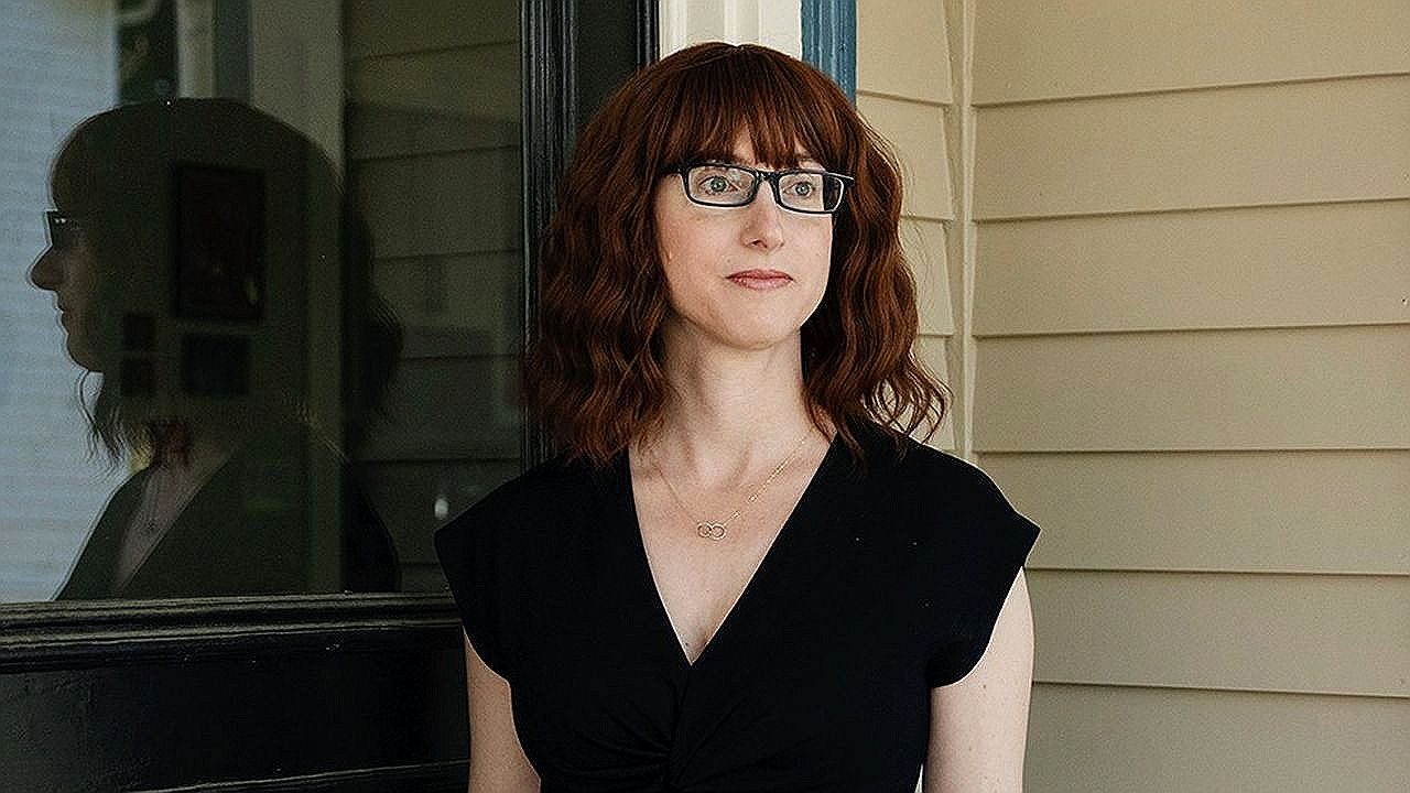 Mulher de cabelo ruivo e óculos observa ao longe em frente a casa em cena de série