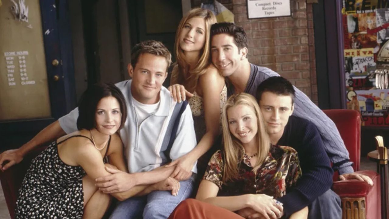 Seis jovens sentados juntos e abraçados em cenário de café com paredes de tijolo posam sorrindo em foto promocional de série ambientada em Nova York