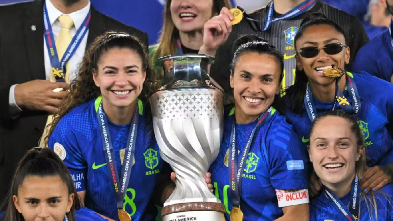 Jogadoras da seleção brasileira de futebol feminino comemoram título segurando um grande troféu prateado; elas vestem uniformes azuis, exibem medalhas e sorriem em celebração coletiva