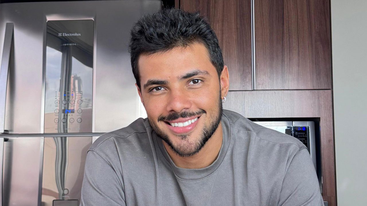 Gabriel Fuentes sorrindo com camiseta cinza em frente a geladeira e armários de cozinha modernos