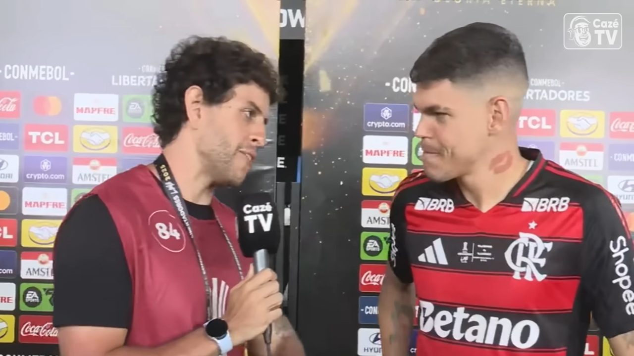 Repórter entrevista um jogador de futebol em frente a um painel repleto de logotipos de patrocinadores da competição. O repórter segura um microfone com a marca da emissora, enquanto o jogador, usando uniforme rubro-negro, ouve atento; ele tem uma marca em forma de beijo no pescoço. O ambiente é típico de zona mista pós-jogo.