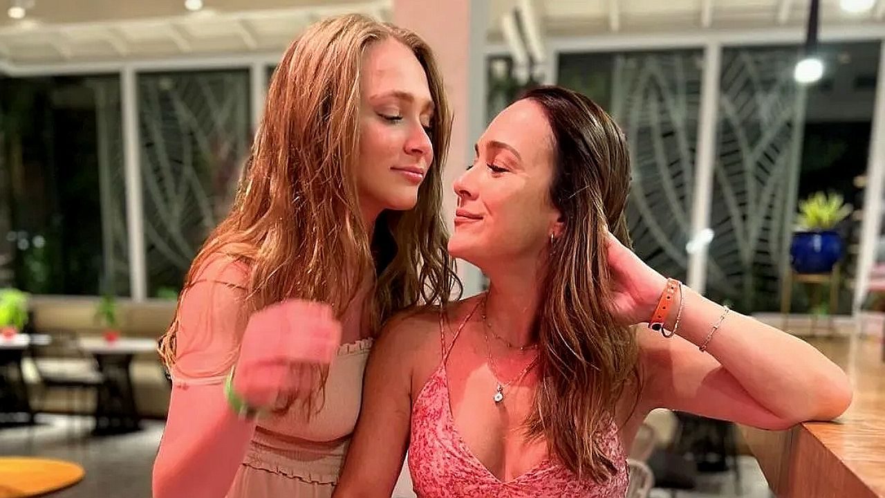 Duas mulheres estão próximas em ambiente interno bem iluminado, apoiadas em um balcão de madeira. Elas se olham com expressões suaves e sorrisos discretos, criando clima de intimidade e carinho. Ao fundo, há janelas amplas e mesas, sugerindo restaurante ou espaço social.