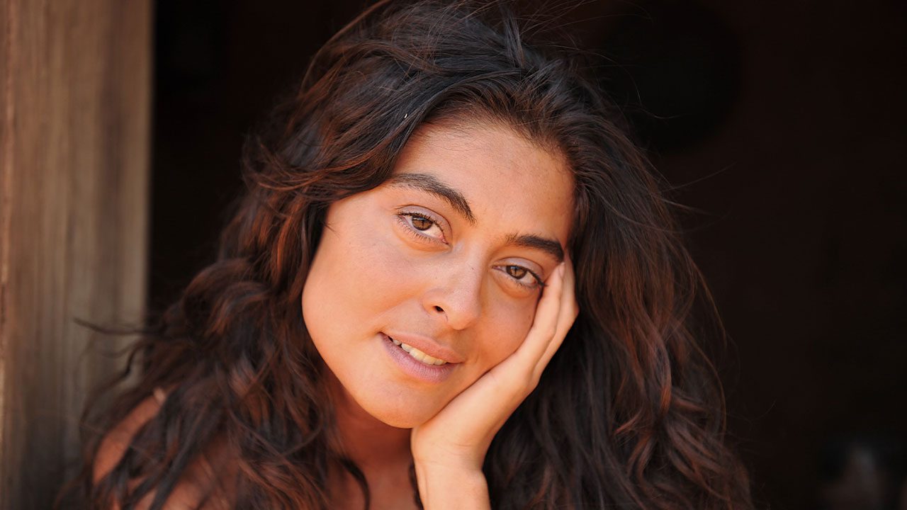 Juliana Paes caracterizada como Gabriela com cabelos soltos apoiando o rosto na mão em ambiente rústico e iluminação natural