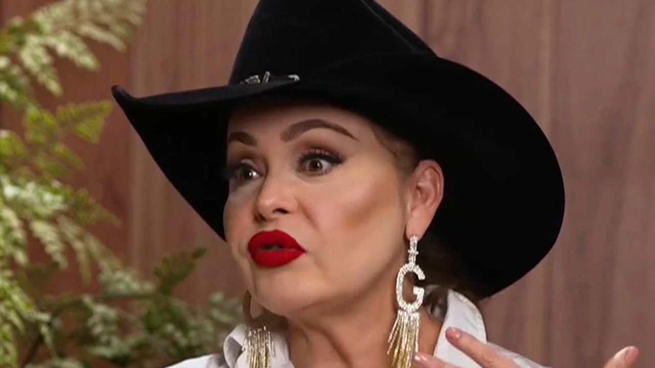 Gaby Spanic usando chapéu preto, brincos longos dourados com a letra G e batom vermelho enquanto fala gesticulando