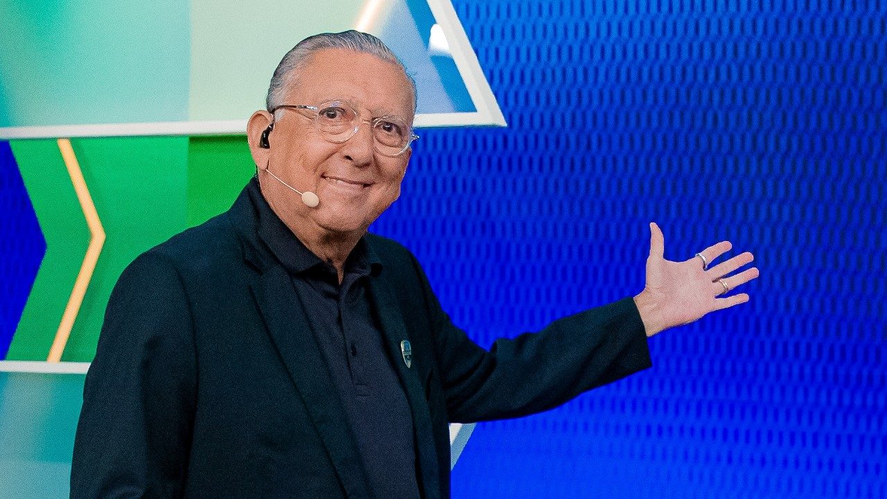 Galvão Bueno sorri e aponta para o cenário azul do programa Galvão F.C. usando microfone de cabeça no SBT