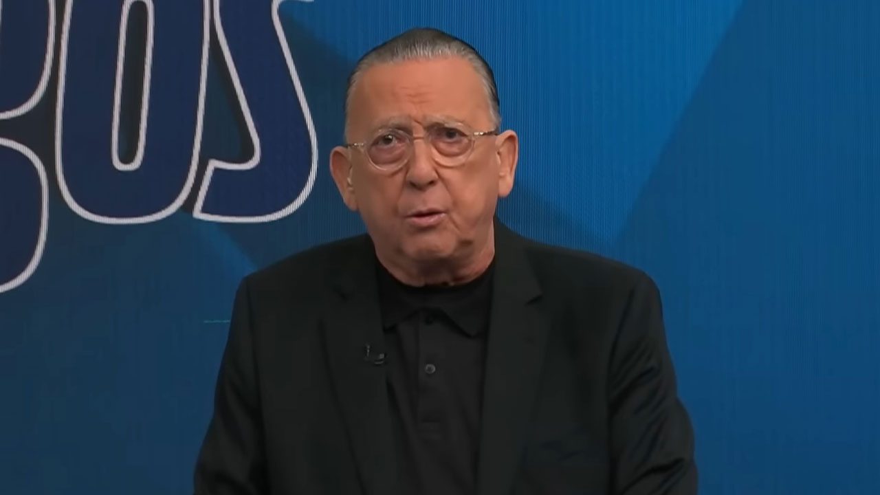 Galvão Bueno usa paletó preto e óculos redondos enquanto fala no cenário azul do programa Galvão e Amigos na Band