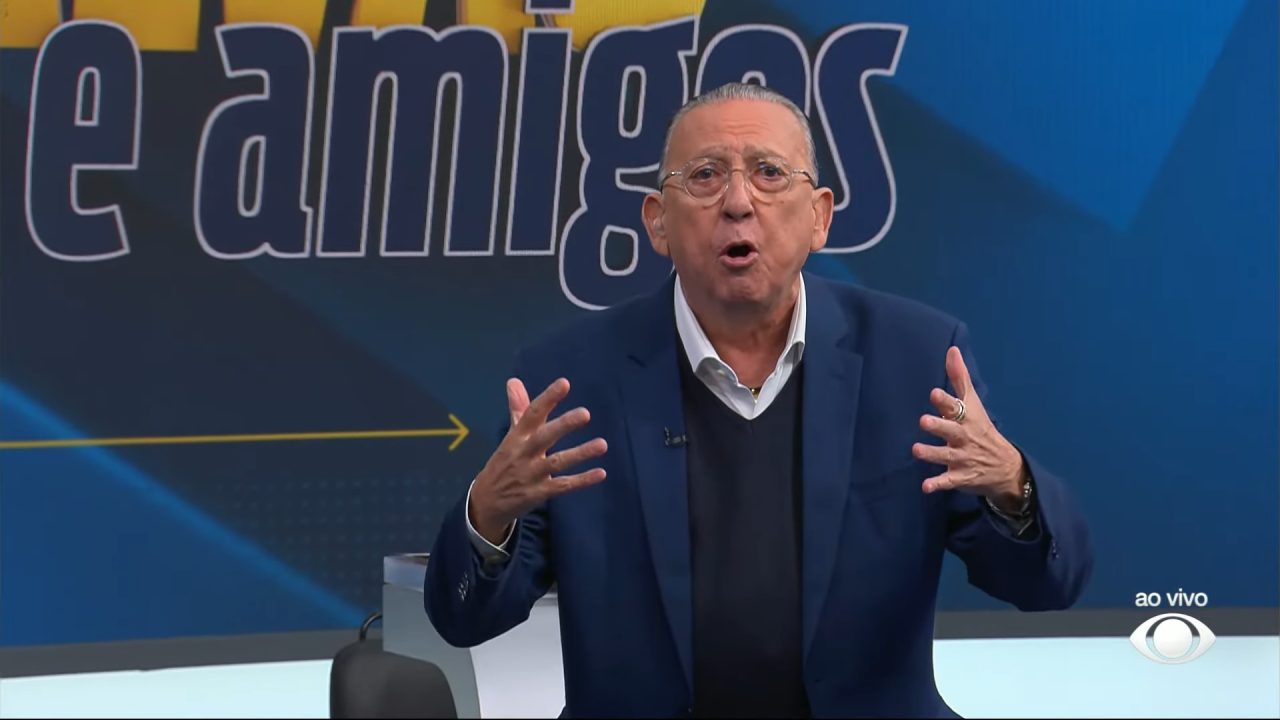 Galvão Bueno, de terno azul e óculos, fala com expressão enfática no estúdio do Galvão e Amigos, gesticulando com as mãos, diante de um painel com letras grandes e coloridas e o logotipo "ao vivo" da Band no canto da tela