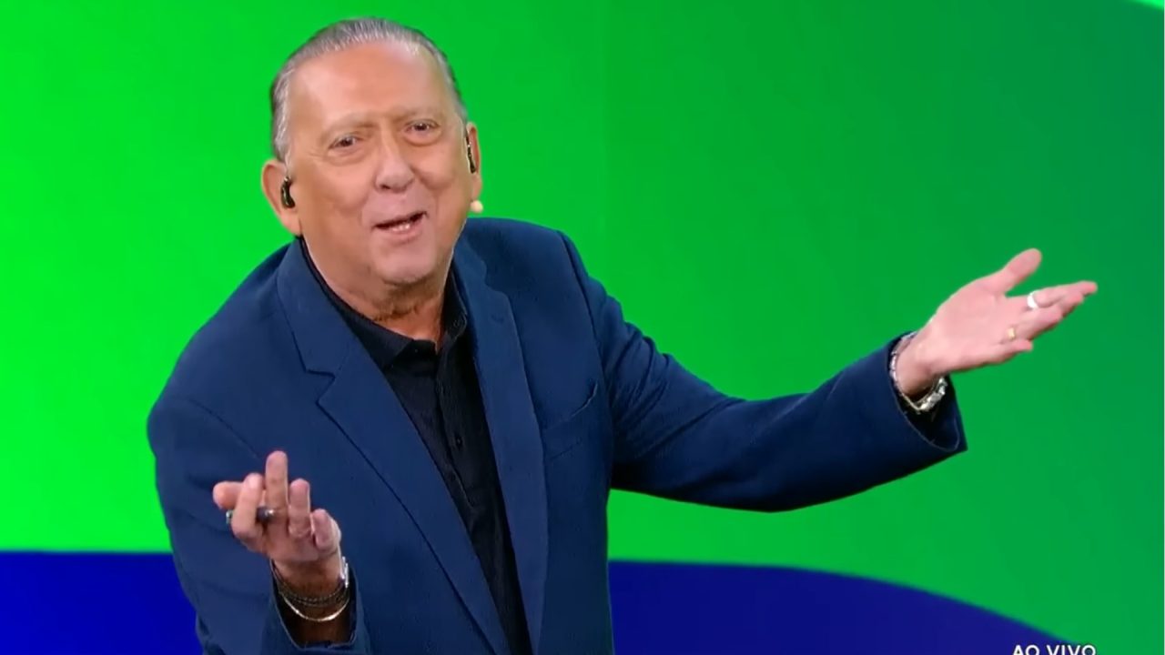 Foto do locutor esportivo Galvão Bueno, vestindo um terno azul, em um cenário moderno do SBT, com fundo verde e azul.