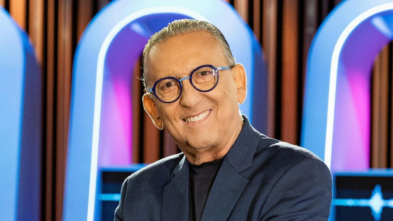 Galvão Bueno sorrindo usa blazer azul escuro e óculos de armação redonda azul em estúdio colorido com luzes de fundo em tons de roxo e azul