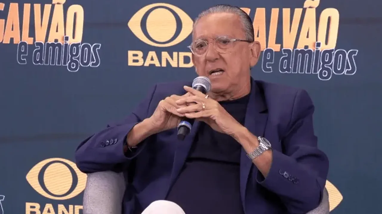 Pessoa idosa sentada em uma poltrona segurando um microfone com as duas mãos, usando óculos de armação fina, blazer escuro sobre camiseta preta e calça clara. A expressão é séria e concentrada, como se estivesse respondendo a uma pergunta em um evento ou entrevista. Ao fundo há um painel com logotipos repetidos, incluindo a palavra “Band” e a frase “Galvão e amigos”, em tons de azul e amarelo, indicando um cenário de programa ou coletiva de imprensa.